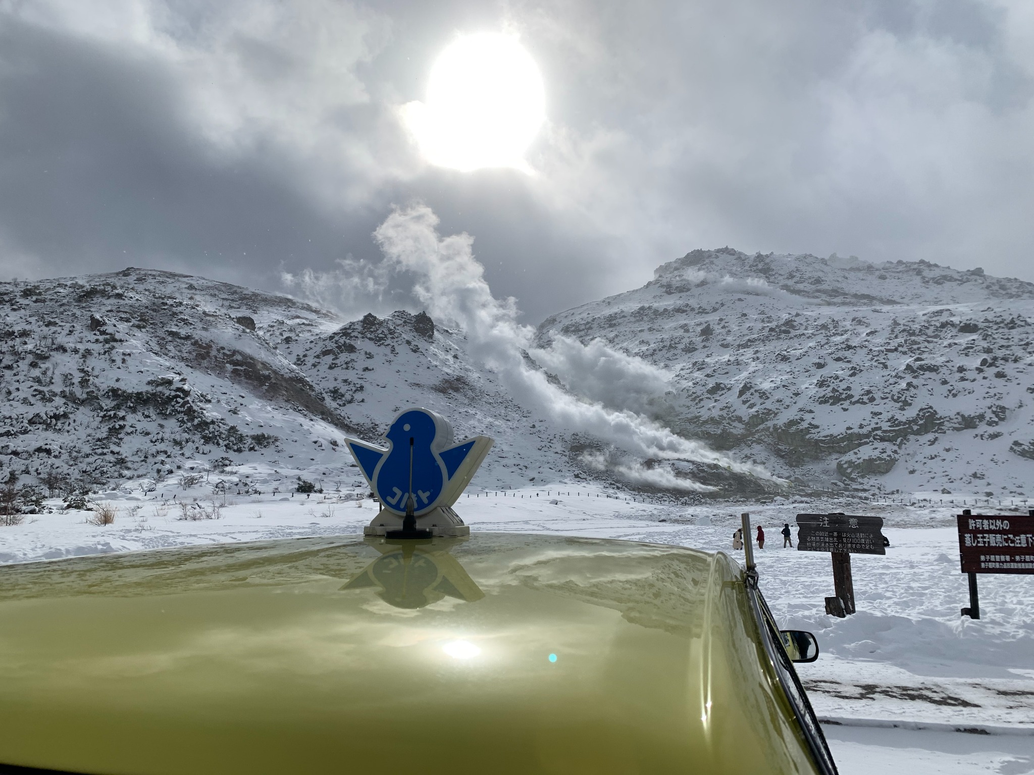 Snowy volcanic landscape with steam, sunlight and a blue bird marker reflected on a car roof.